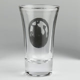 A clear glass shot glass featuring a pewter Viking ship medallion on the front. Shown from the back