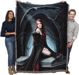 A large woven throw blanket with fringe edges is held up by a man and a woman against a plain white background. The blanket features a gothic illustration of a vampire woman with long red hair, surrounded by bats in a dark blue and black archway scene.