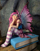 Fairy figurine with pink hair and wings sitting on a blue book against a stone wall background.