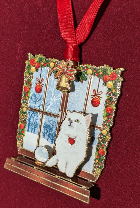 Brass ornament of a white cat sitting in a festive windowsill with snow beyond and a snow globe.
