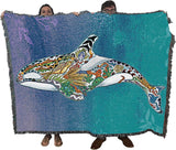 A large woven throw blanket with fringe edges is held up by two people against a white background. The blanket features a colorful, patterned orca whale design over a blue and teal gradient background.