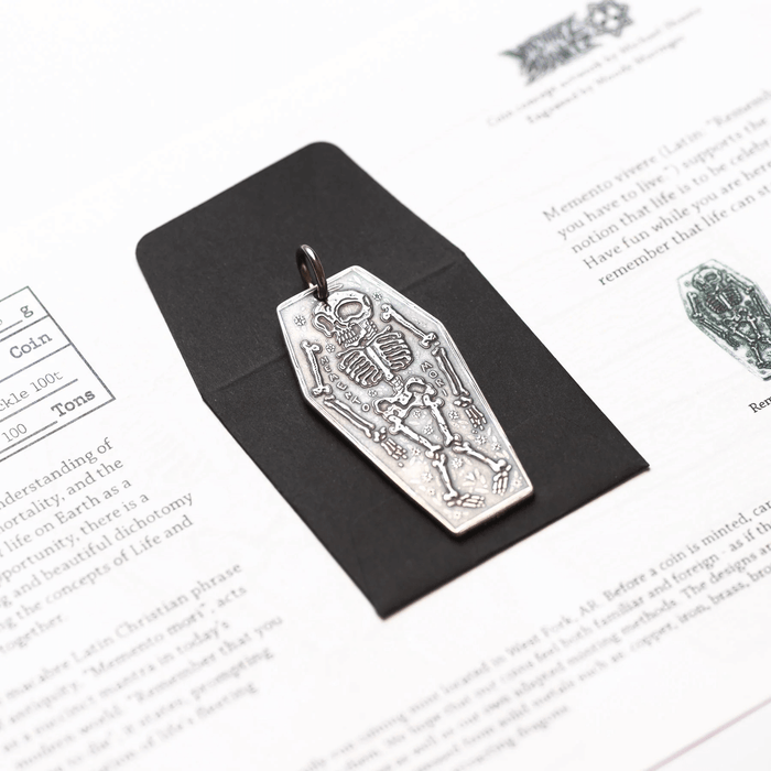 Silver charm featuring a skeleton and the phrase "Memento Mori", presented with envelope and information