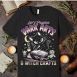 Black t-shirt with a cat graphic and text 'Let's Practice Dark Arts & Witch Crafts' on a rustic background.