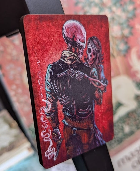 Magnet showing skeleton couple embracing, each holding a gun, with red background