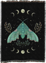 A black woven throw blanket with fringe edges, featuring a large teal-green moth surrounded by cream-colored crescent moons, stars, and botanical motifs.