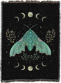 A black woven throw blanket with fringe edges, featuring a large teal-green moth surrounded by cream-colored crescent moons, stars, and botanical motifs.