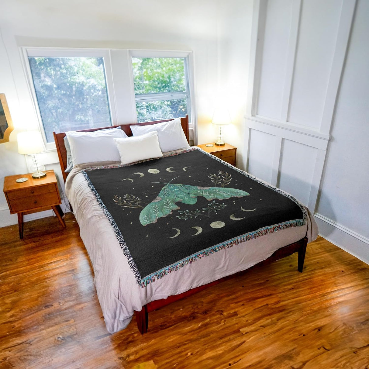 A black quilted blanket featuring a large teal-green moth and moon pattern, styled on a bed in a bright bedroom with wood floors and bedside tables.