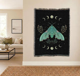 A black woven wall tapestry with a large teal moth and moon motifs, hanging on a beige wall in a modern living room setting.