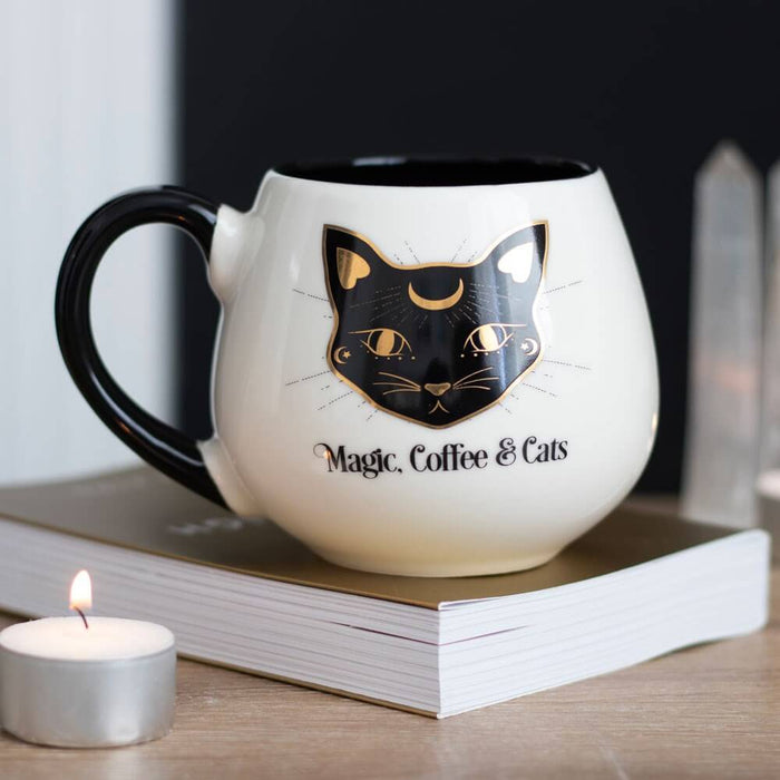 Magic, Coffee & Cats mug in white, black and gold with cat face, shown sitting on a book near a tealight