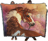 A large woven throw blanket with fringe edges featuring a detailed illustration of chestnut pinto horse, held up by two people whose heads and feet are visible behind the blanket against a white background.