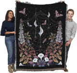 Two people holding a decorative blanket with spider web and moth design against a black background
