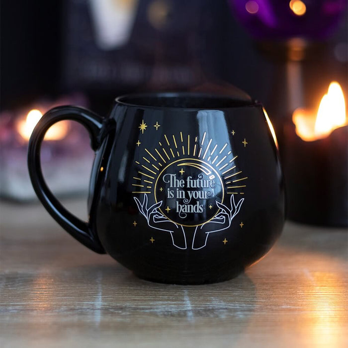 Fortune Teller Color Change mug. Black base with hands and a crystal ball. The words "The future is in your hands" are revealed when a hot beverage is poured in. Shown on a table with firelight in the background