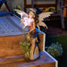 Solar powered fairy figurine shows of a pixie in pink admiring tall flowers. Shown with the blossoms glowing at night, displayed on a wooden deck
