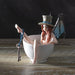 Fairy with steampunk wings in blue and a tophat, and a key looped around her leg. She sits in a teacup, taking a bath. Shown displayed on a wooden table