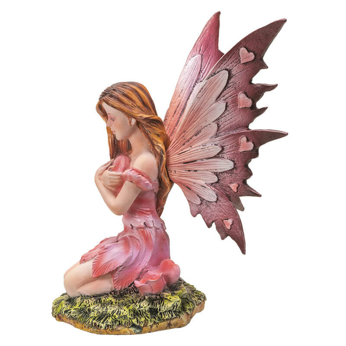 Fairy with pink dress and wings, with hearts on wings. She holds a heart and sits next to rose flowers on the grass. Shown from the side