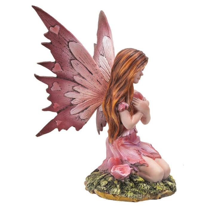 Fairy with pink dress and wings, with hearts on wings. She holds a heart and sits next to rose flowers on the grass. Shown from the side