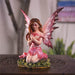 Fairy with pink dress and wings, with hearts on wings. She holds a heart and sits next to rose flowers on the grass. Shown on a wooden table