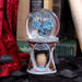 Waterglobe snowglobe with two blue dragons facing each other inside. Ornate base with golden dragon designs and a bronze dragon egg at the center. Displayed in front of leather bound books and a red velvet curtain with black flower petals.