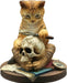 Figurine of an orange tabby cat holding a magnifying glass up to a human skull. The kitty sits amidst books, maps and a compass. 