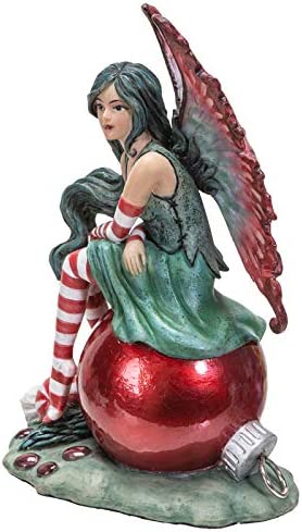 Fairy sitting on a red Christmas ornament. She wears a green dress with red & white striped stockings and sleeves and there is a peppermint nearby. Side view