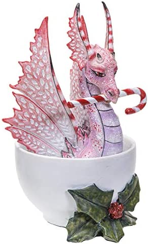 Pink and red dragon sitting in a cup, holding a candy cane in its mouth. Holly leaves and berries sit at the base of the cup. Side view