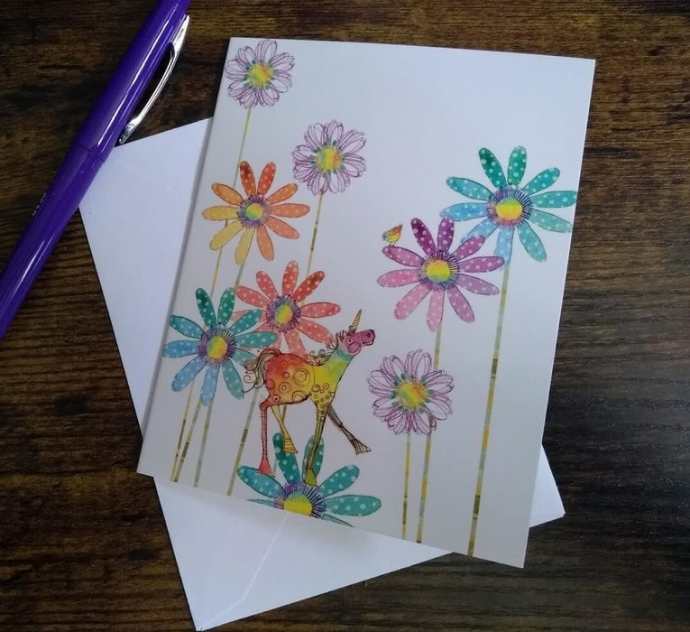 Unicorn notecard showing a rainbow unicorn on a polka dot blue flower, with more blooms around in other colors. Sitting on top of its white envelope, with a purple pen nearby, ready to be written