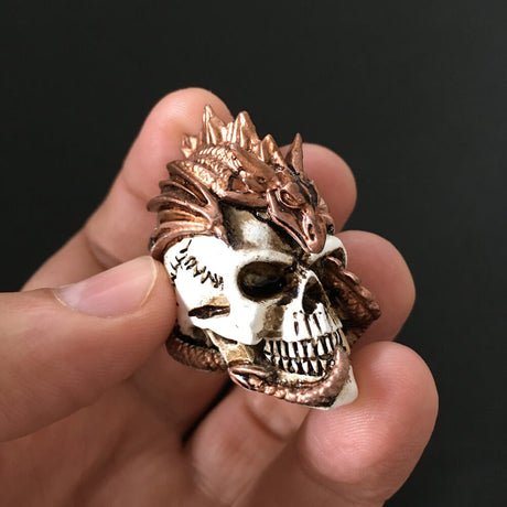 Miniature figurine of a bronze dragon curling around and through a human skull. Shown held in a hand, it is small