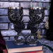 Black Cat goblet pair. Each wine glass has a black cat with purple eyes on the stem and silver swirls on the cup, with a stainless steel insert. Displayed on a stack of books in front of a brick wall
