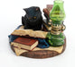 Black cat reading a book next to more tomes and a green lamp. Shown head-on, cat has golden orange eyes and sits on a blue cloth
