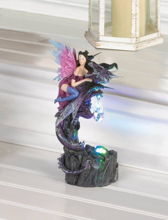 Figurine showing a dragon with pink and blue wings in a purple dress and indigo stockings. She holds onto a purple dragon who has a crystal dangling from its claws. The figure is glowing with an LED and placed on a white shelf