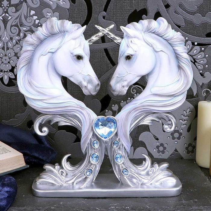 Figurine with two unicorn busts facing one another, silver horns crossed. Unicorns are done in shades of white and pale silver, blue and purple. A light cyan heart gem and three oval blue gems adorn the front of the base. Displayed in front of a black and gray ornate swirling backdrop