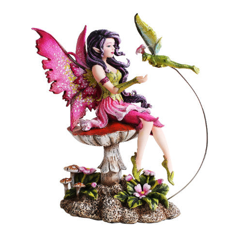 A fairy in pink and green greets her pixie winged frog prince. The fae sits on a mushroom amidst flowers
