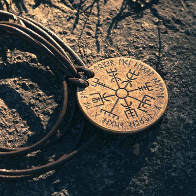 Necklace pendant version of the Vegvesir Bronze Norse Compass coin showing runes, on leather cord shown on a rock in the sun