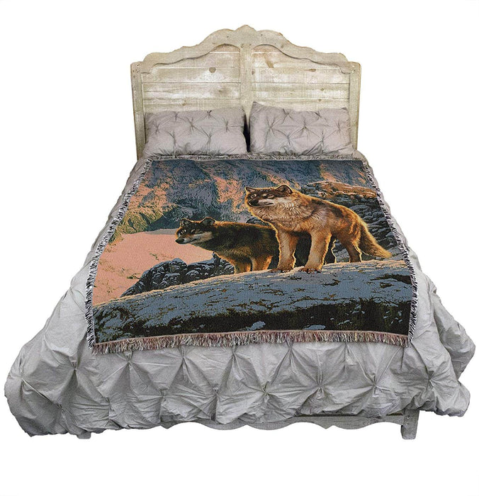 Tapestry blanket showing two wolves standing in the sunset, in a winter mountain landscape. Shown draped over a bed